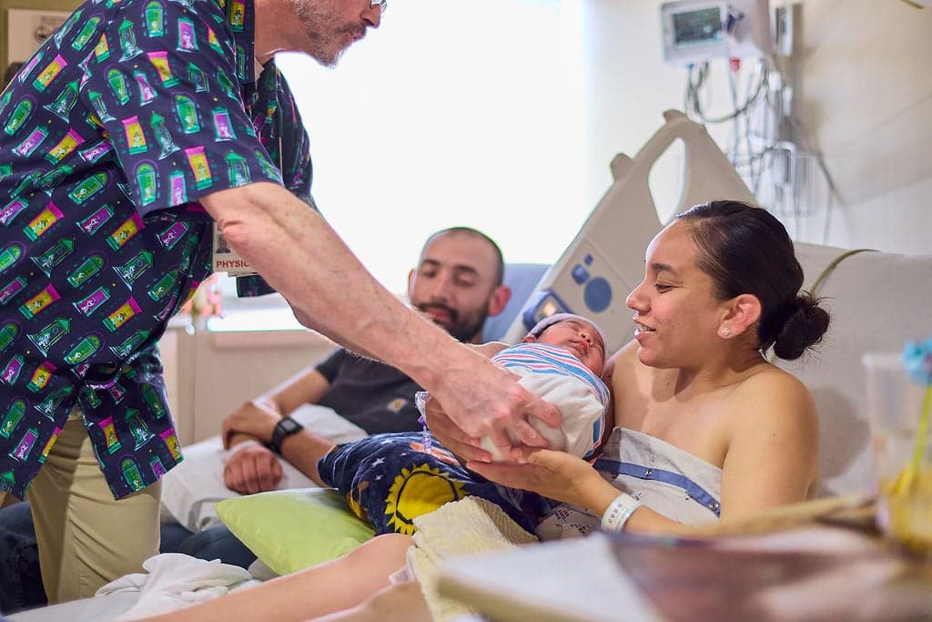 An ob-gyn hands a newborn baby to a new mother in her hospital bed.