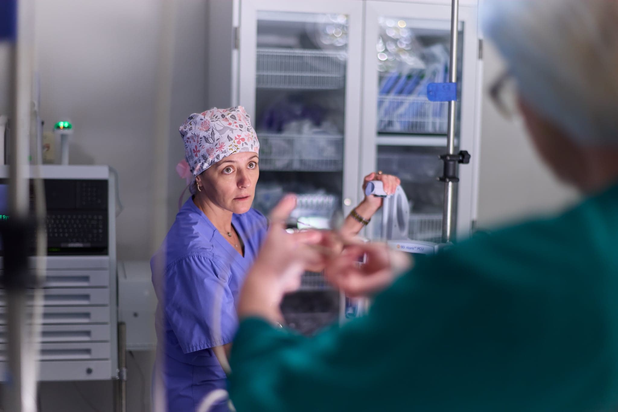 A surgical team member in a scrub cap talks with a colleague in an operating room.