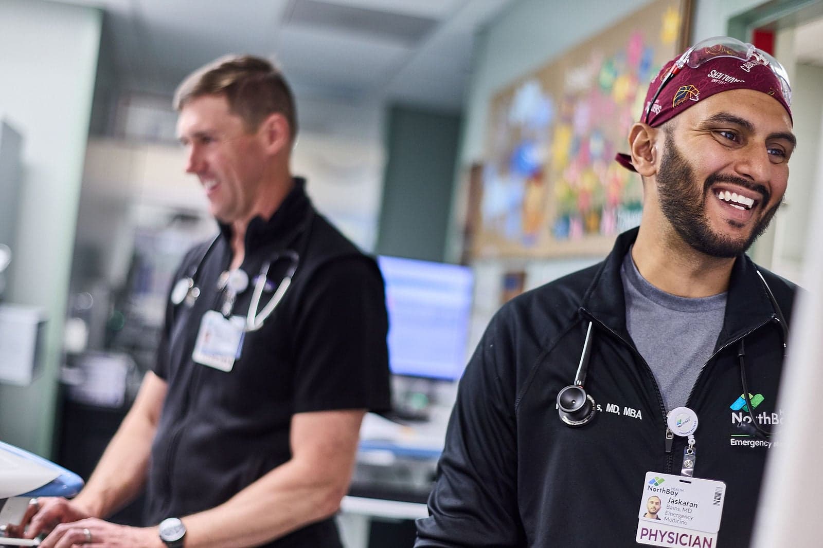 Two doctors smile while looking at their computers in an emergency room.