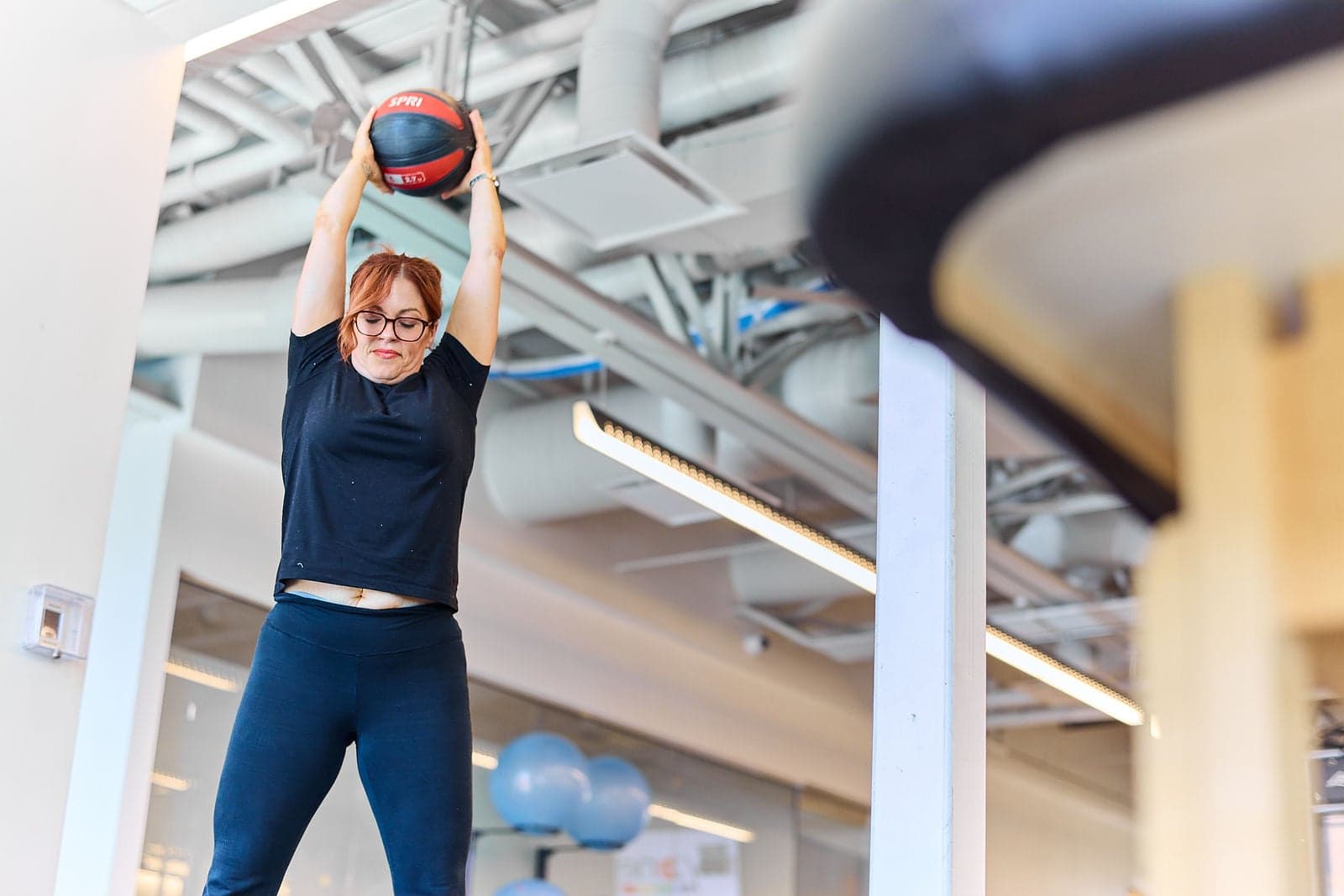 A woman lifts a weighted ball overhead in a rehabilitation gym.