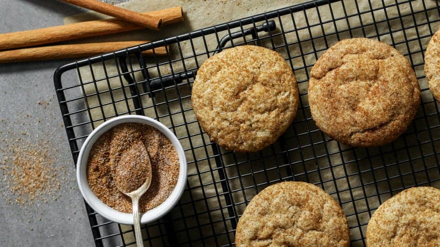 Some snickerdoodles cooling on a black wire rack with a bowl of cinnamon sugar next to them on the counter.
