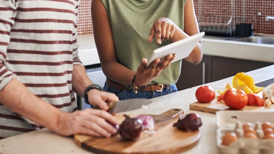 A young couple in the kitchen working on preparing a meal while going over the recipe on their phone.