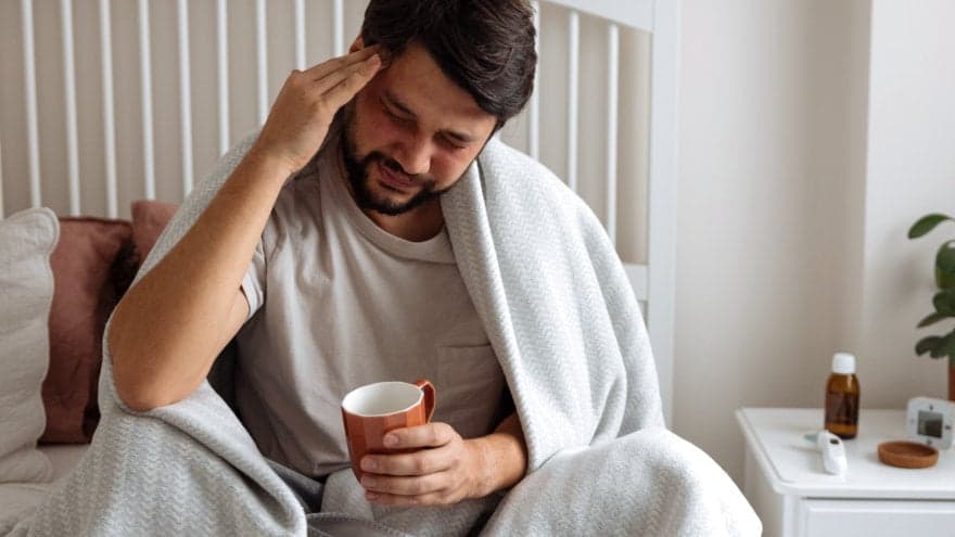 A man with his eyes closed, rubbing his pained head while holding a tea mug in bed. On his bedside table medicine and a thermometer can be seen.
