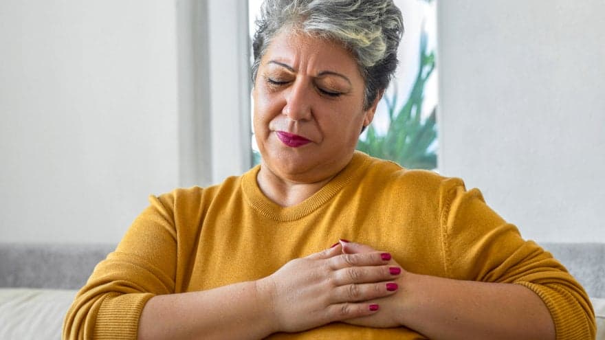 A mature woman in her 40-50s with pixie-length grey hair and a mustard yellow sweater. Her eyes are closed, with her face slightly scrunched and hands pressing against her chest in a sign of pain.