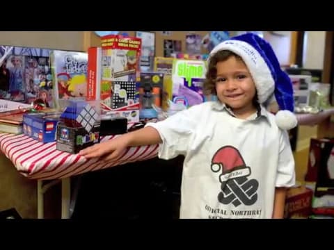 Child wearing festive Santa hat and graphic t-shirt posing with holiday-themed toy display on striped table