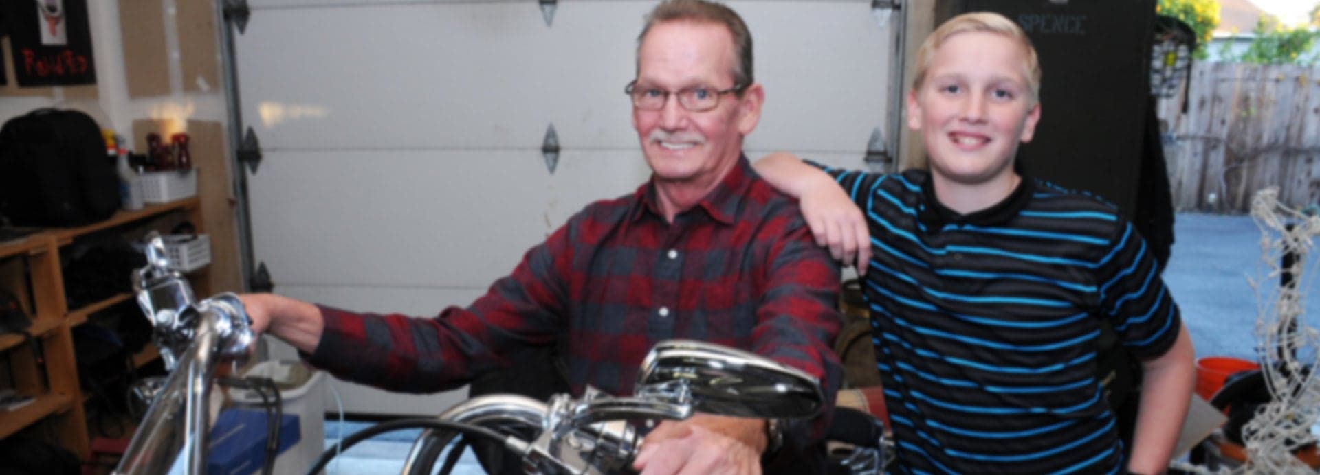 Two men smiling in a garage, one wearing a red plaid shirt with his arm around the other in a blue striped shirt, standing be
