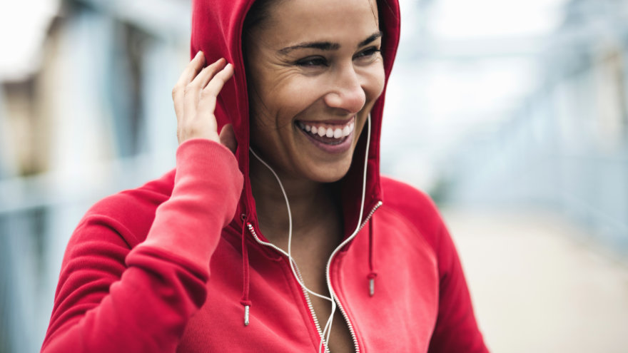 Woman in a red zipper jacket looking away from the camera and smiling as she adjusts her in-ear headphones before her workout.