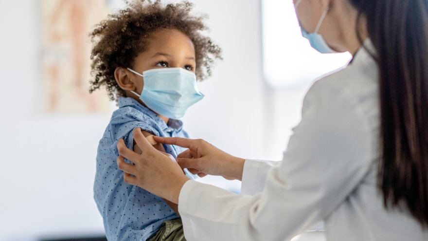 Small child looking up at his doctor who just gave him a shot and is putting a bandage over the area. They are both wearing blue surgical masks.