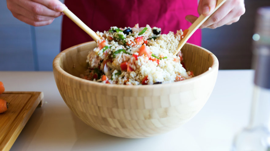 A man wearing a burgundy red apron tossing together quinoa salad with wooden spoons in a wooden bowl.