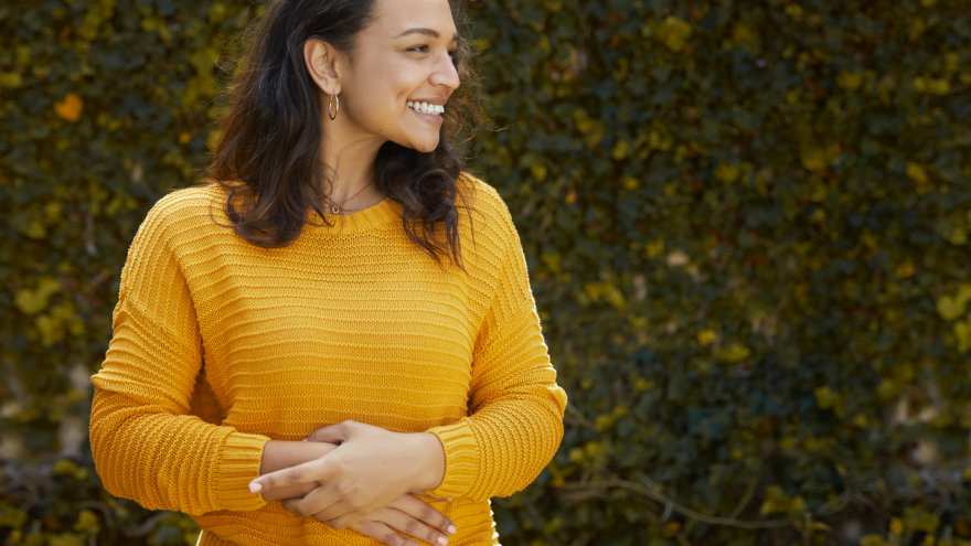 A woman wearing a cozy orange sweater standing in front of a ivy covered wall. She has her hands overlapped on her stomach and is looking to the left, smiling.