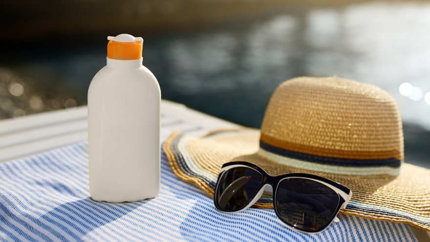 A bottle of sunscreen, a wide-brimmed straw hat and some sunglasses sitting on top of a striped blue towel on a slatted white bench. Shimmering water can be seen in the background.