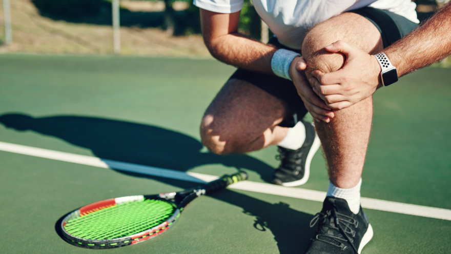 A man taking a break from playing tennis to crotch down and hold his knee.