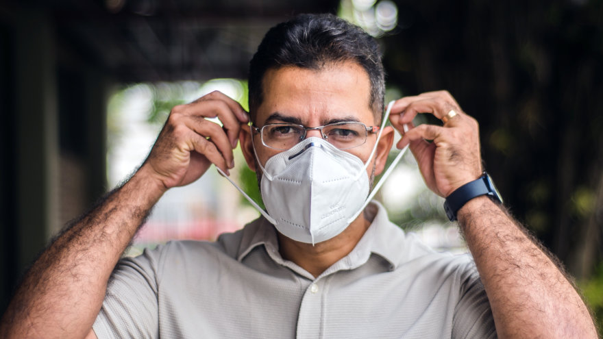 Man with short-cropped, black hair outside facing the camera while placing a N95 mask on.