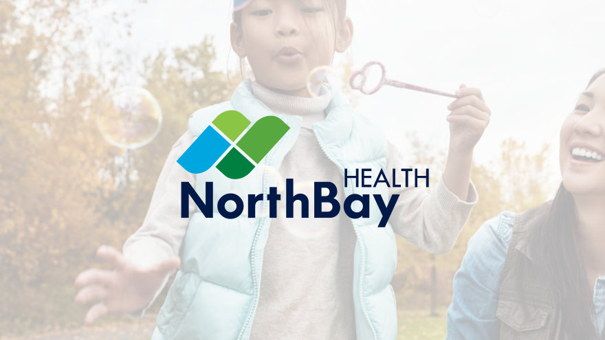 A little girl blowing bubbles towards the camera while her mother looks on from the side, laughing. The new NorthBay Health logo appears in the middle on a transparent white overlay.