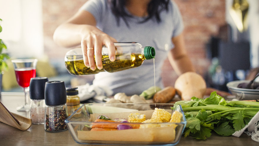 A woman drizzling oil over vegetables she's preparing to roast. Spices and a glass of wine can be seen in the background.