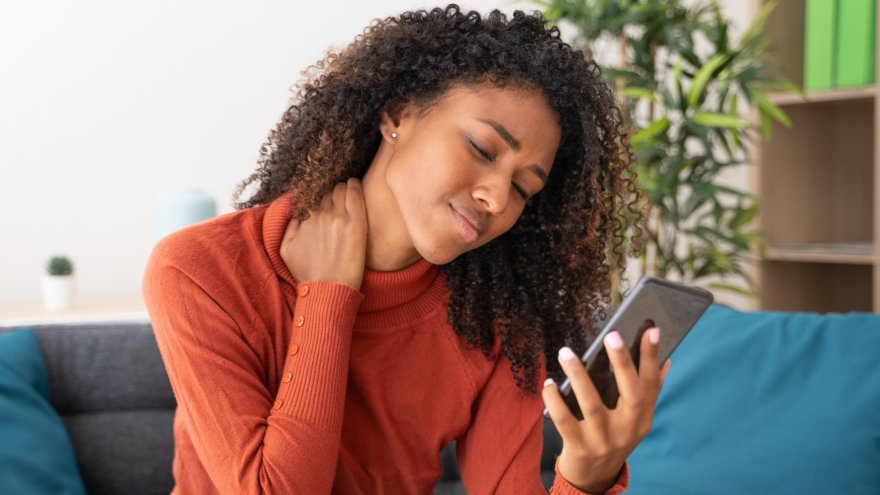A girl rubbing her neck from pain while looking down at her phone.