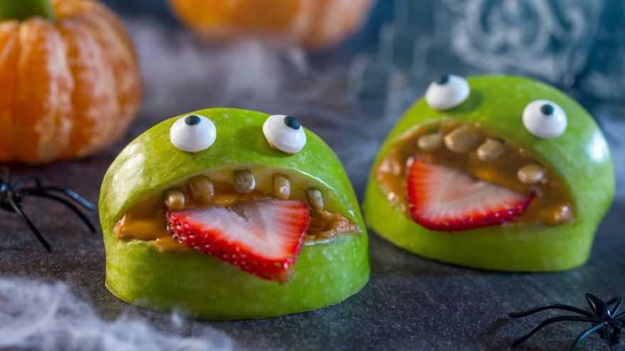 Two apple slices arranged to look like an open mouth, with peanut butter spread on the inside to hold pumpkin seeds as teeth, and a strawberry slice for a tongue. Two edible eyes are placed on the top apple slice.