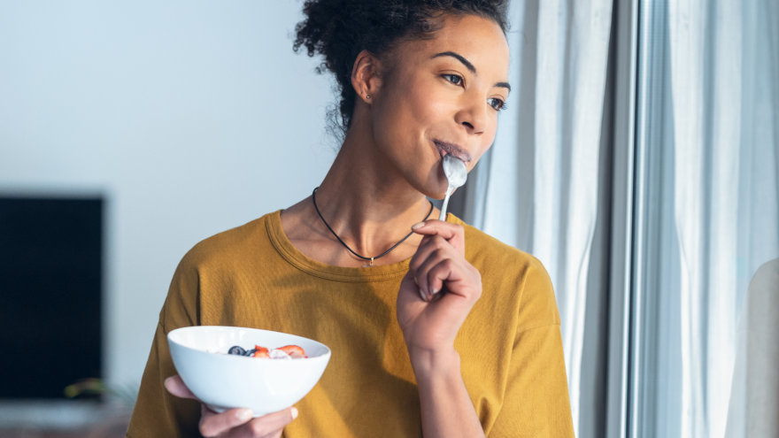 A woman enjoying a bowl of yogurt and fruit, the spoon still in her mouth as she gazes out her window.