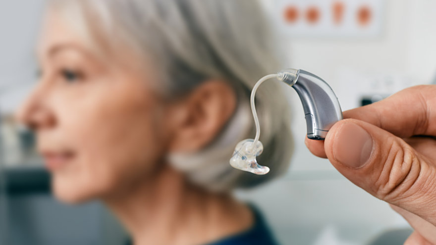 A sleek, modern hearing device is held up in the foreground. The profile of a mature woman appears blurred in the background that looks like an audiologist's office.