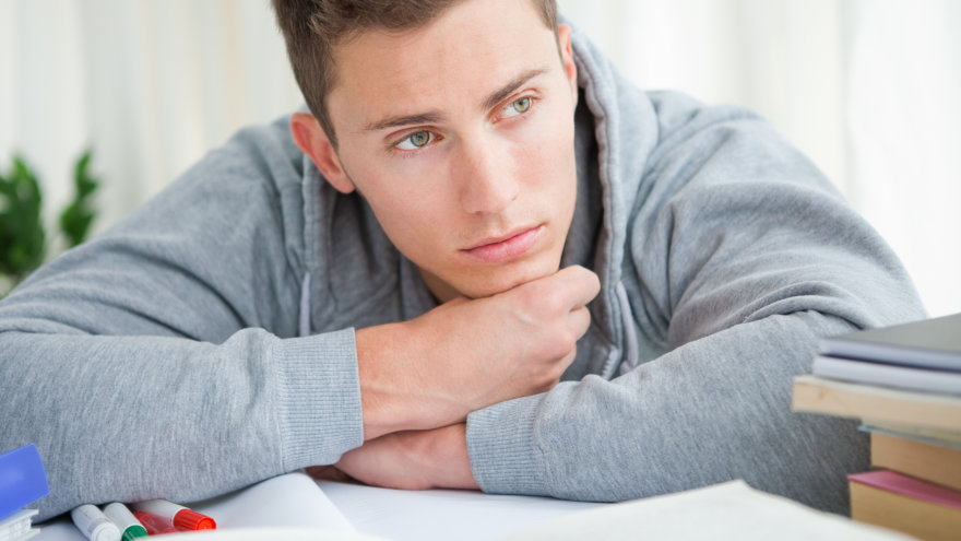 Young white male student taking a break, leaning forward while resting on his desk. He looks away from the camera with a distracted, worried expression.