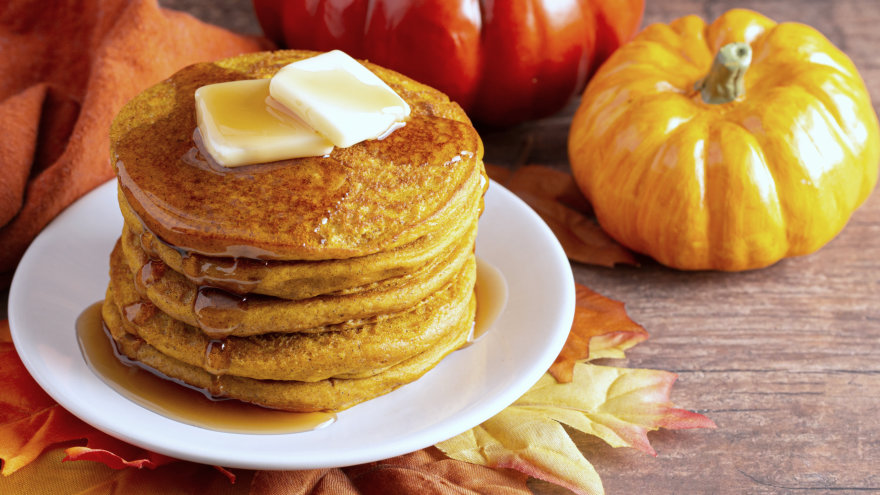 Stack of pumpkin pancakes on a plate, surrounding by small pumpkins and yellow and orange maple leaves.