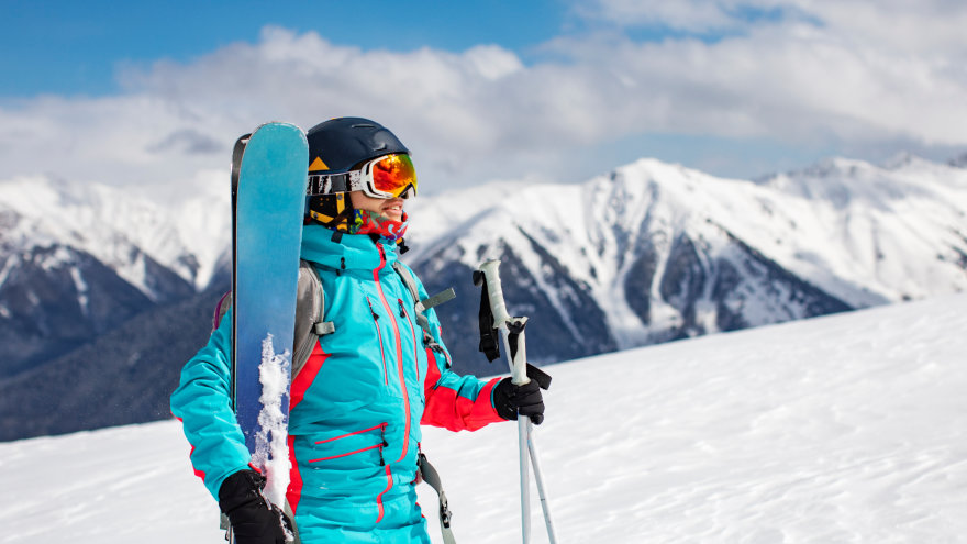 A woman bundled in full skiing attire, holding her snow covered skis and ski poles as she walks on the slope with blue skies and snow covered mountains in the background.
