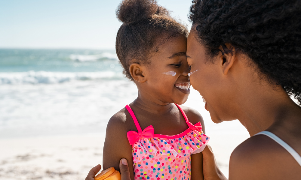 Mother playfully touching her nose to her daughter's. A bottle of sunscreen can be seen in the mother's hand, and some on the cheeks of the daughter. The waves and sandy shore of the beach are behind them.