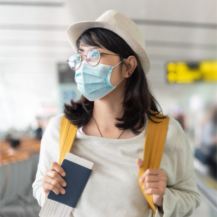 Woman at the airport with a mask and passport in hand, holding onto the straps of her yellow backpack.