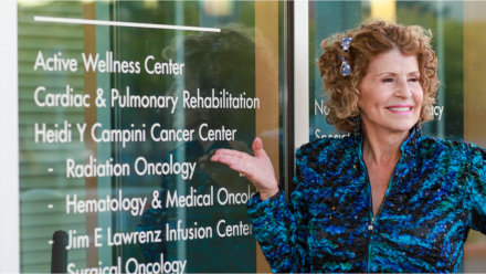 Heidi Y Campini standing in front of the updated Cancer Center entrance, highlighting the door's vinyl lettering reflecting the new name.